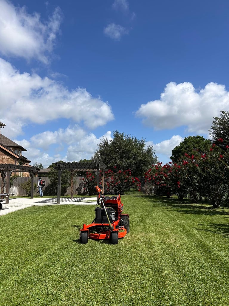 Red zero-turn mower on green lawn with garden, flowering trees, and pergola under blue sky with white clouds