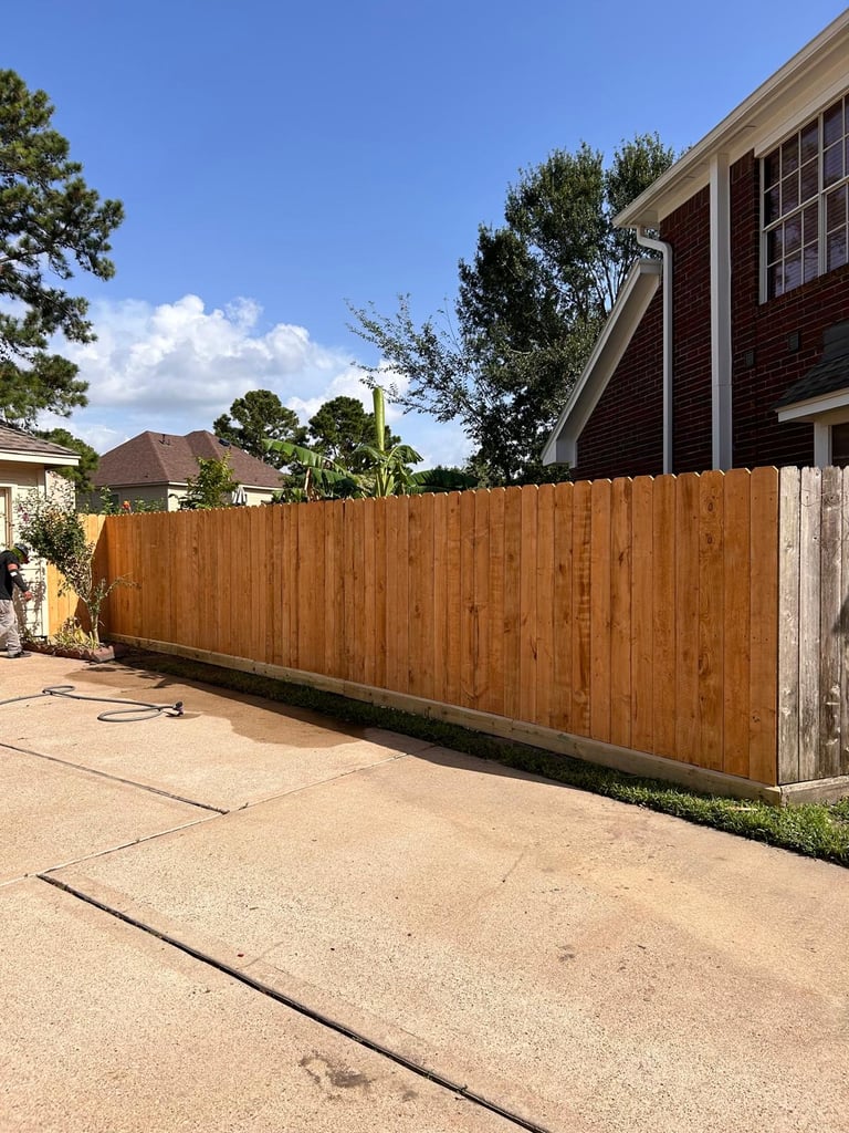 Brown wooden fence along a residential driveway with a brick house and trees in the background under blue sky