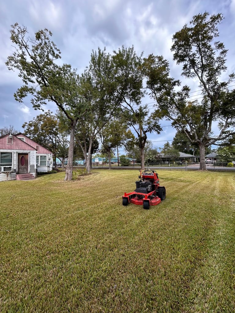 Red riding mower on large green lawn with pink cottage and tall trees in background